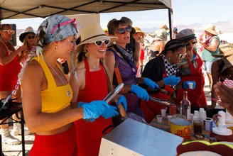 A group of smiling people wearing blue gloves prepare and serve hot dogs and cold drinks to participants in Black Rock City