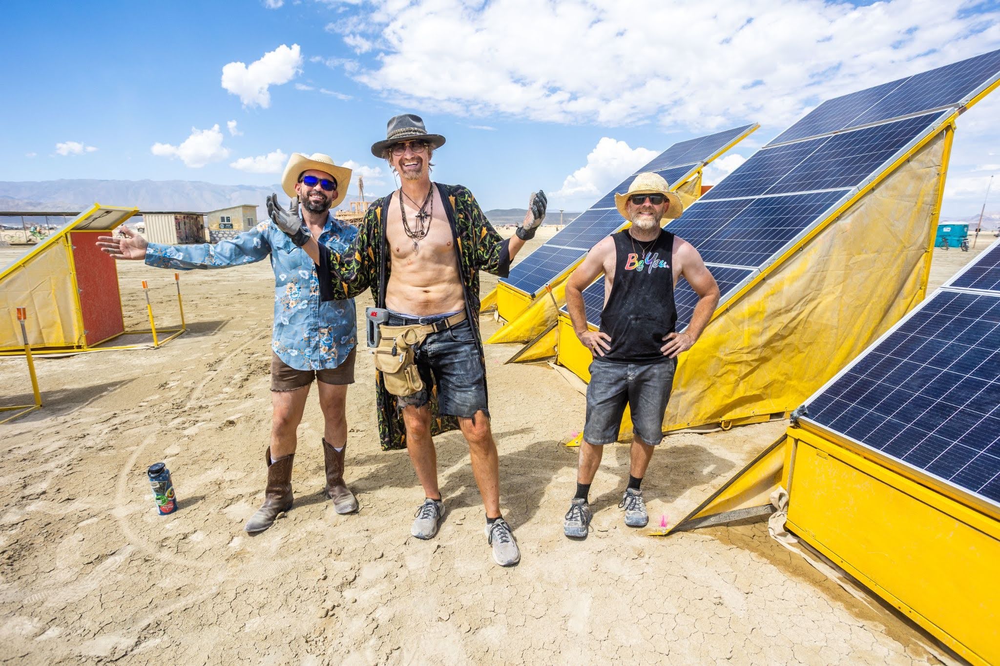 Three people stand next to solar arrays in Black Rock City arms open welcomingly.
