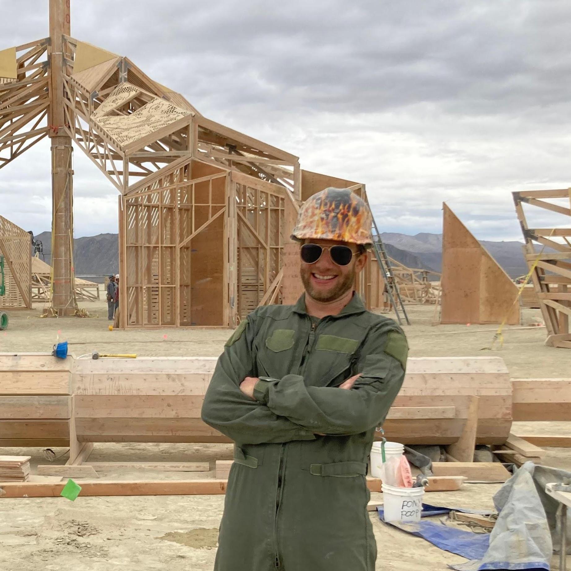 Steve wearing yellow hardhat and green jumpsuit in front of the construction site.